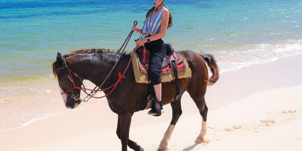 Scenic horse riding experience on the beach, with riders trotting along the shoreline, the gentle waves lapping at their feet, and the sun setting in the background, creating a tranquil and picturesque coastal adventure.